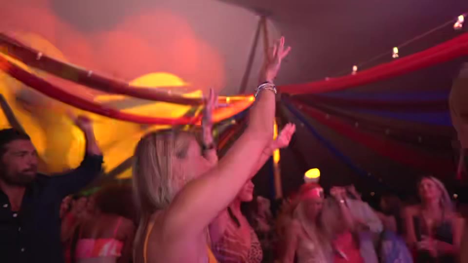 A packed floor under coloured tent drapes and string lights, hands in the air.