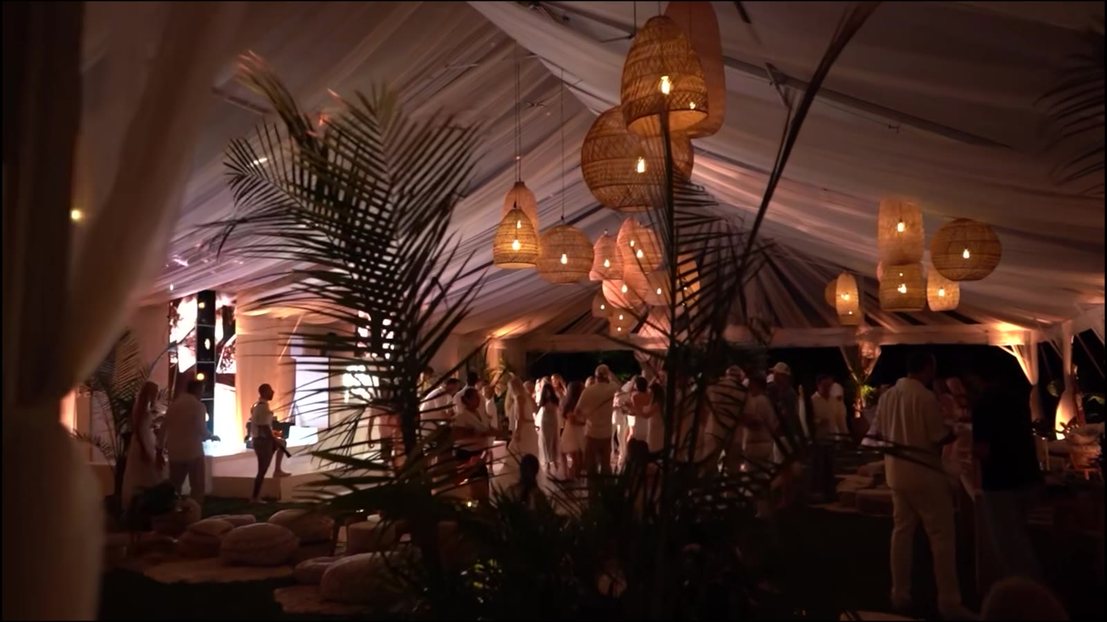 A tented room at night: woven rattan pendants hung over palms, with a crowd dressed in white on the floor beneath them.